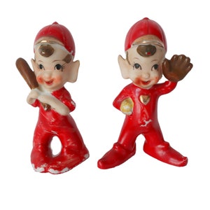 May include: Two red and white ceramic elf figurines wearing baseball caps. One elf is holding a baseball bat and the other is holding a baseball glove.
