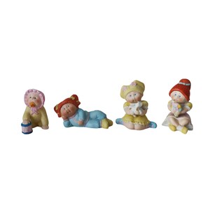 May include: Four small, colourful figurines of babies. One baby is wearing a pink bonnet and holding a drum. Another is lying down in blue pyjamas. The other two are sitting, one holding a toy and the other holding flowers.