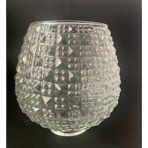 May include: A clear glass bowl or light fixture shade featuring a repeating geometric pattern of raised squares and triangular facets. The object has a rounded, bulbous shape with a smooth circular opening at the top and a narrow base.