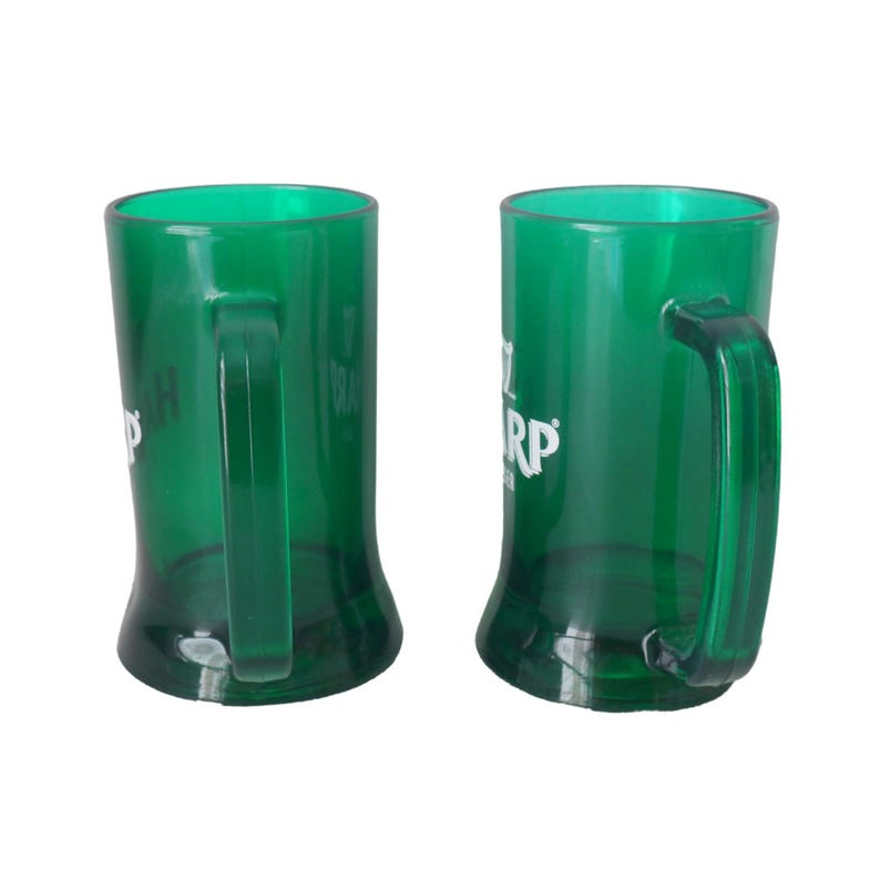 May include: Two green beer tankards with handles. The tankards have a curved shape and a clear, transparent appearance. The tankards have white text on them. The tankards are empty and sitting on a white surface.