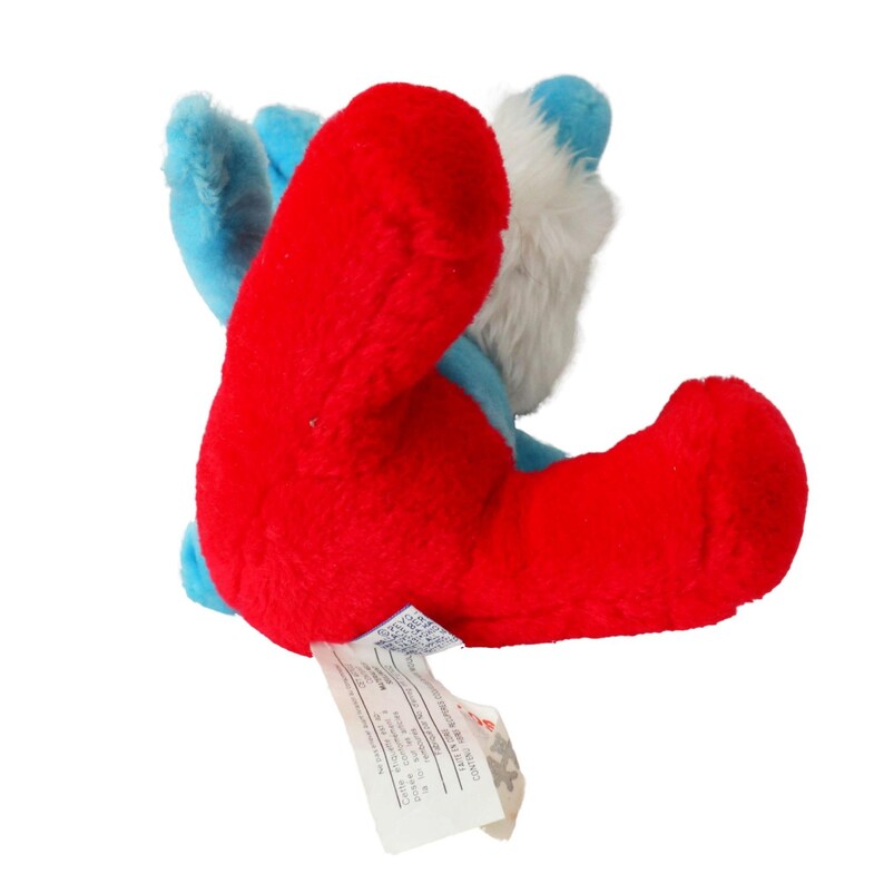 May include: A red and blue plush toy with a white face. The toy is lying on its back with its legs in the air. The toy has a tag attached to it that says "Made in China".