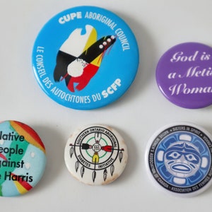 May include: A collection of five round buttons with various designs and text. One button is blue with text and a graphic, another purple with "God is a Metis Woman", and others with text and graphics related to Indigenous issues.