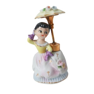 May include: A vintage porcelain figurine of a woman holding a parasol. The figurine is dressed in a yellow and white dress with pink accents and gold detailing. The parasol is cream-colored with floral accents.