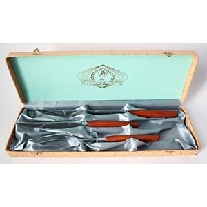 May include: A vintage cutlery set in a decorative box. The set includes knives and serving utensils with wooden handles. The box is lined with satin and features a logo that reads "The Connoisseur's Choice."