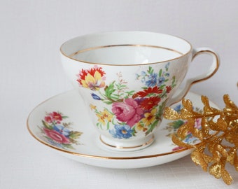 May include: A white teacup and saucer with a floral pattern in shades of pink, red, yellow, and blue. The cup and saucer have a gold trim.