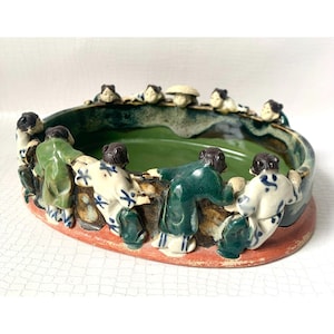 May include: A ceramic bowl with a green interior and a red base. The bowl is decorated with small figures in green and white clothing, appearing to hold hands around the rim. The figures are in a circle, with some figures looking over the edge.