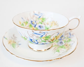 May include: A white teacup and saucer with a floral pattern featuring blue and yellow flowers. The cup and saucer have a gold trim.