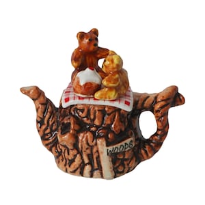 Puede incluir: Una caprichosa tetera de cerámica marrón con un diseño texturizado similar a la corteza. Dos pequeños ositos de peluche están sentados encima de una tela a cuadros rojos y blancos, junto con una magdalena. La tetera tiene un asa y un pico, con la palabra "WOODS" en un pequeño letrero.
