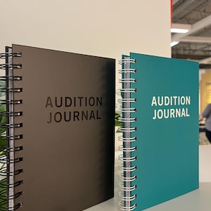 May include: Two spiral-bound journals, one brown and one teal, are displayed. The text "AUDITION JOURNAL" is printed on the front of each journal in a sans-serif font. The journals are on a white surface.