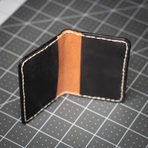 Puede incluir: Cartera de cuero negro con interior marrón y costuras blancas. La cartera plegable está abierta, mostrando los colores y texturas contrastantes del cuero. La cartera está hecha a mano y tiene un diseño minimalista.