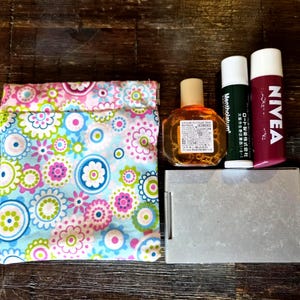 May include: A floral print pouch in blue, pink, and green, next to a small bottle, two lip balms, and a silver compact. The pouch has a repeating pattern of flowers and circles. The items are arranged on a dark wood surface.