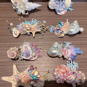 Op de afbeelding: Een verzameling handgemaakte haarclips met schelpen, parels en decoratieve elementen. De clips tonen een verscheidenheid aan kleuren, waaronder wit, roze, blauw en iriserende accenten. Elke clip heeft een uniek ontwerp, met zeester- en schelpvormen.