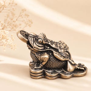 May include: A bronze-colored metal figurine of a three-legged toad sitting on a pile of coins. The toad has a textured, bumpy back and holds a single coin with Chinese characters in its mouth, while additional coins are visible on its back.