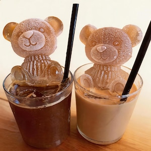 Könnte beinhalten: Zwei Gläser, gefüllt mit Eiskaffee und Eiskaffee Latte, jeweils mit einem eiswürfelförmigen Eisbären. Die Eisbären haben detaillierte Merkmale und sitzen in den Getränken. Schwarze Strohhalme sind in jedem Glas platziert.