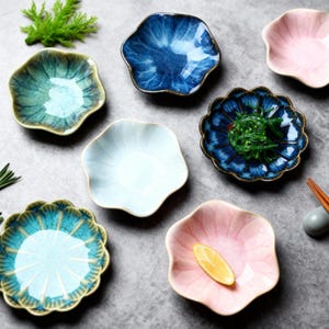 May include: A collection of ceramic, petal-shaped dishes in various colors, including blue, pink, and turquoise. Some dishes contain food, such as seaweed and a lemon slice. The dishes have a textured, glazed finish and are arranged on a gray surface.