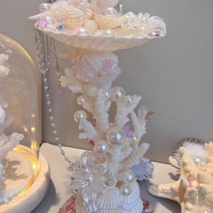 May include: A decorative seashell centerpiece featuring a white coral-like structure adorned with pearls and various seashells. The centerpiece includes a large, open seashell bowl at the top, with a pearl necklace hanging down.