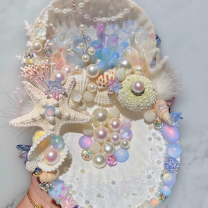 Könnte beinhalten: Ein dekoratives Muschelkunstwerk mit einer Vielzahl von Elementen. Es zeigt einen Seestern, Perlen, Muscheln und schillernde Edelsteine. Die Farbpalette ist überwiegend weiß, mit Akzenten in Rosa, Blau und Lila. Das Stück ist mit kleinen Perlen und Kristallen verziert.