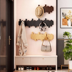 Op de afbeelding: Een moderne entree met drie houten kapstokken in zwart, bruin en goud. Een strohoed, koptelefoon en een handtas hangen aan de kapstokken. Een bank met schoenenopslag staat eronder, met zichtbare schoenen.
