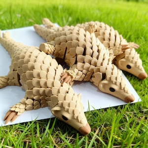 May include: Three tan pangolin sculptures are displayed on a white surface, with green grass in the background. The sculptures feature a detailed, scale-like texture. The pangolins are positioned to suggest movement.