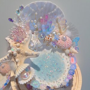 Peut inclure: Présentoir décoratif sur le thème des coquillages, avec une zone centrale bleu clair pailletée, entourée de coquillages, d'étoiles de mer, de perles et de cristaux irisés. Le présentoir est monté sur une base en bois, créant une esthétique fantaisiste inspirée de l'océan.