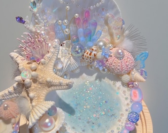 May include: A decorative seashell-themed display featuring a central light blue glittered area, surrounded by seashells, starfish, pearls, and iridescent crystals. The display is mounted on a wooden base, creating a whimsical, ocean-inspired aesthetic.
