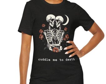May include: Black t-shirt featuring a graphic of two skeletons embracing, surrounded by red roses and a crescent moon. The text "cuddle me to death" is printed below the graphic. The shirt is made of a soft material.