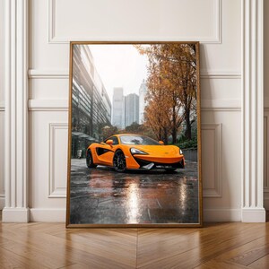 Könnte beinhalten: Gerahmter Druck eines orangefarbenen Sportwagens, der auf einer nassen Stadtstraße geparkt ist. Das Auto, mit seinem schlanken Design und den schwarzen Rädern, steht im Mittelpunkt. Der Hintergrund zeigt Gebäude und Bäume mit Herbstlaub.