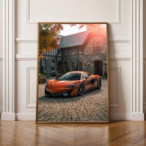 Könnte beinhalten: Gerahmter Druck mit einem orangefarbenen Sportwagen, der auf einer Kopfsteinpflasterauffahrt vor einem Steingebäude geparkt ist. Das Auto hat ein schlankes Design. Das Bild ist in einem Holzrahmen gerahmt.