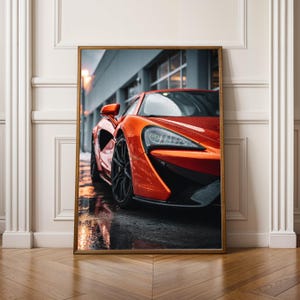 Könnte beinhalten: Gerahmter Druck eines leuchtend orangefarbenen Sportwagens. Das schlanke Design des Autos wird durch seine schwarzen Räder und die nasse Fahrbahn hervorgehoben. Das Bild ist vor einem verschwommenen urbanen Hintergrund platziert, was ein Gefühl von Geschwindigkeit und Luxus erzeugt.