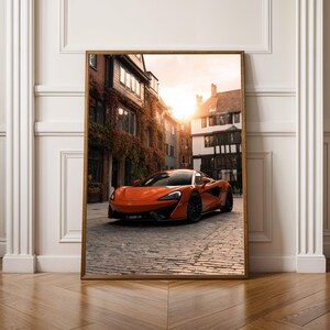 Könnte beinhalten: Gerahmter Druck eines orangefarbenen Sportwagens auf einer Kopfsteinpflasterstraße. Das Auto steht im Mittelpunkt, vor dem Hintergrund europäischer Architektur und eines warmen Sonnenuntergangs. Der Druck wird auf einem Holzboden präsentiert.