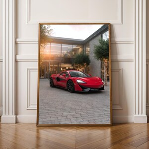 Könnte beinhalten: Gerahmter Druck eines roten Sportwagens auf einer gepflasterten Auffahrt. Das Auto hat ein schlankes, modernes Design mit schwarzen Rädern. Im Hintergrund befindet sich ein Gebäude mit großen Fenstern und Bäumen.
