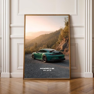 Peut inclure: Impression encadrée d'une Porsche 911 GT3 verte sur une route de montagne. La voiture est sur un chemin de gravier avec une forêt et un coucher de soleil en arrière-plan. L'impression est dans un cadre en bois.