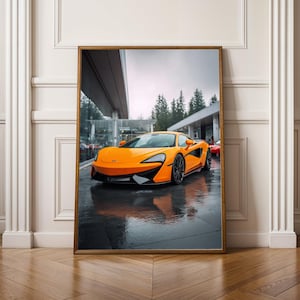 Könnte beinhalten: Gerahmter Druck eines orangefarbenen Sportwagens, der auf einer nassen Oberfläche geparkt ist. Das Auto hat schwarze Räder und Akzente. Der Hintergrund umfasst ein Gebäude mit Glasfenstern und Bäumen. Das Bild wird in einem Raum mit Holzboden und weißer Wand angezeigt.