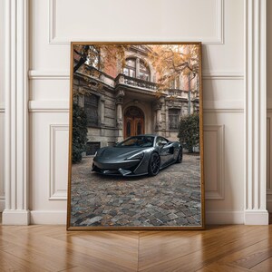 Könnte beinhalten: Gerahmter Druck eines grauen Sportwagens, der auf einer Kopfsteinpflasterauffahrt vor einem stattlichen Gebäude geparkt ist. Das Auto hat ein schlankes Design mit schwarzen Rädern. Das Gebäude hat einen klassischen architektonischen Stil mit Bogenfenstern und einer Holztür.