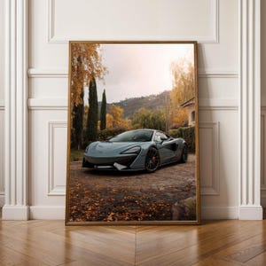 Könnte beinhalten: Gerahmter Druck eines grauen Sportwagens auf einer Backsteinfahrt. Das Auto steht vor herbstlichen Bäumen und einem Haus. Der Druck wird auf einem Holzboden präsentiert.