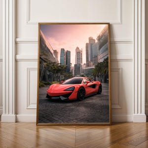 Könnte beinhalten: Gerahmter Druck mit einem leuchtend orangefarbenen Sportwagen auf einer Kopfsteinpflasterstraße. Der Hintergrund zeigt eine moderne Stadtlandschaft mit hohen Gebäuden und einem Sonnenuntergangshimmel. Der Rahmen ist aus Holz.