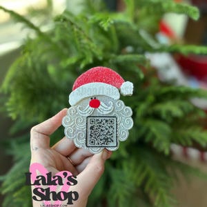 May include: A Santa Claus-shaped ornament with a red hat, white beard, and a QR code. The ornament has a glittery silver finish and a red nose. The text "Lala's Shop" and the website address are visible in the image. The background is a blurred Christmas tree.