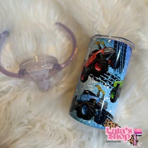 May include: A stainless steel tumbler with a lid and a clear sippy cup top. The tumbler features a blue background with illustrations of red, blue, and green monster trucks. The text "Lala's Shop" is visible.