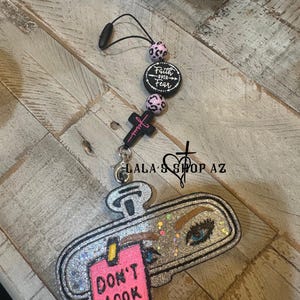 May include: A car charm featuring a rearview mirror design with a pink "Don't Look Back" tag. The charm includes a cross with "Jesus" and beads with a leopard print pattern. The charm is silver and black.