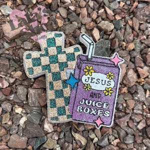 May include: Glittery cross and juice box-shaped pins. The cross has a green and gold checkered pattern. The purple juice box reads "JESUS AND JUICE BOXES" with yellow flower and pink star accents. The pins are on a rocky surface.