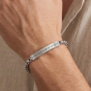 May include: A silver bracelet with a rectangular plate engraved with coordinates. The bracelet features a chain-link design with small, rounded beads. The coordinates are 25° 47 16.32 N, 80° 07 39.24 W.