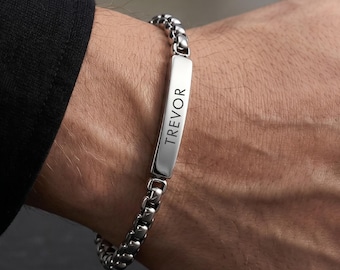 May include: A silver-colored bracelet with the name "TREVOR" engraved on a rectangular plate. The bracelet has a chain-link design and is worn on a wrist.