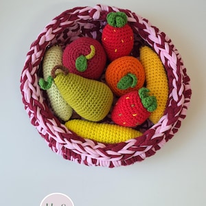 May include: A round, hand-crocheted basket in alternating shades of dark burgundy and light pink, filled with a collection of crocheted play food items including a green pear, a red apple, two strawberries, an orange, and two yellow corn cobs.