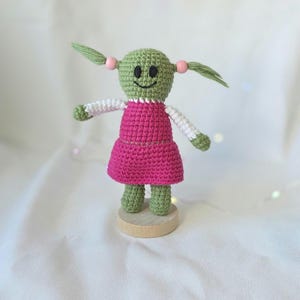 May include: A handmade crochet doll with a green head and pigtails, wearing a pink dress and white sleeves. The doll has a smiling face and is standing on a small wooden base. The doll is approximately 5 inches tall.