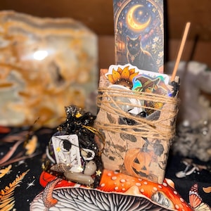 May include: A rectangular paper package wrapped in twine, featuring a pumpkin and floral print, topped with a bookmark showing a black cat under a crescent moon. Beside it sits a black organza drawstring bag containing small crystals and a tarot card.