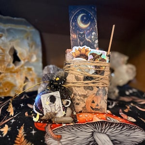 May include: A rectangular package wrapped in brown paper with a pumpkin and leaf print, tied with twine, containing stickers of a sunflower and a witch. A black organza drawstring bag with a tarot card and a small shell sits beside the package.