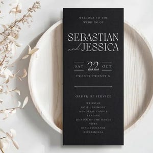 Può includere: Un programma di nozze nero con testo bianco, esposto su un piatto di legno chiaro. Il programma recita "Benvenuti al matrimonio di Sebastian e Jessica" con la data "22 OTT Twenty Twenty X" e l'ordine della cerimonia.