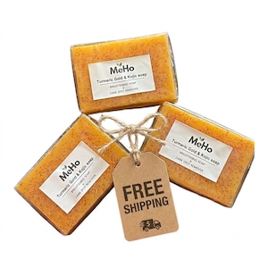 May include: Three rectangular bars of orange turmeric gold and kojic soap with visible dark specks, each wrapped in clear plastic with a white label reading MeHo Turmeric Gold & Kojic soap, tied together with twine and a brown paper tag.