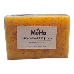 May include: A rectangular bar of orange soap with small dark brown specks throughout. The label reads MeHo Turmeric Gold & Kojic soap, BRIGHTENING SOAP + DARK SPOT REMOVER. The soap is wrapped in clear plastic film, showing a smooth, solid texture.