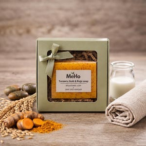 May include: A rectangular bar of orange turmeric and kojic soap with visible dark specks, packaged in a light green gift box with a clear window and a matching ribbon bow. The label reads MeHo Turmeric Gold & Kojic soap BRIGHTENING SOAP + DARK SPOT REMOVER.